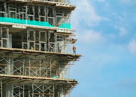 A construction worker stands on a scaffolding at a high-rise building under a blue sky.